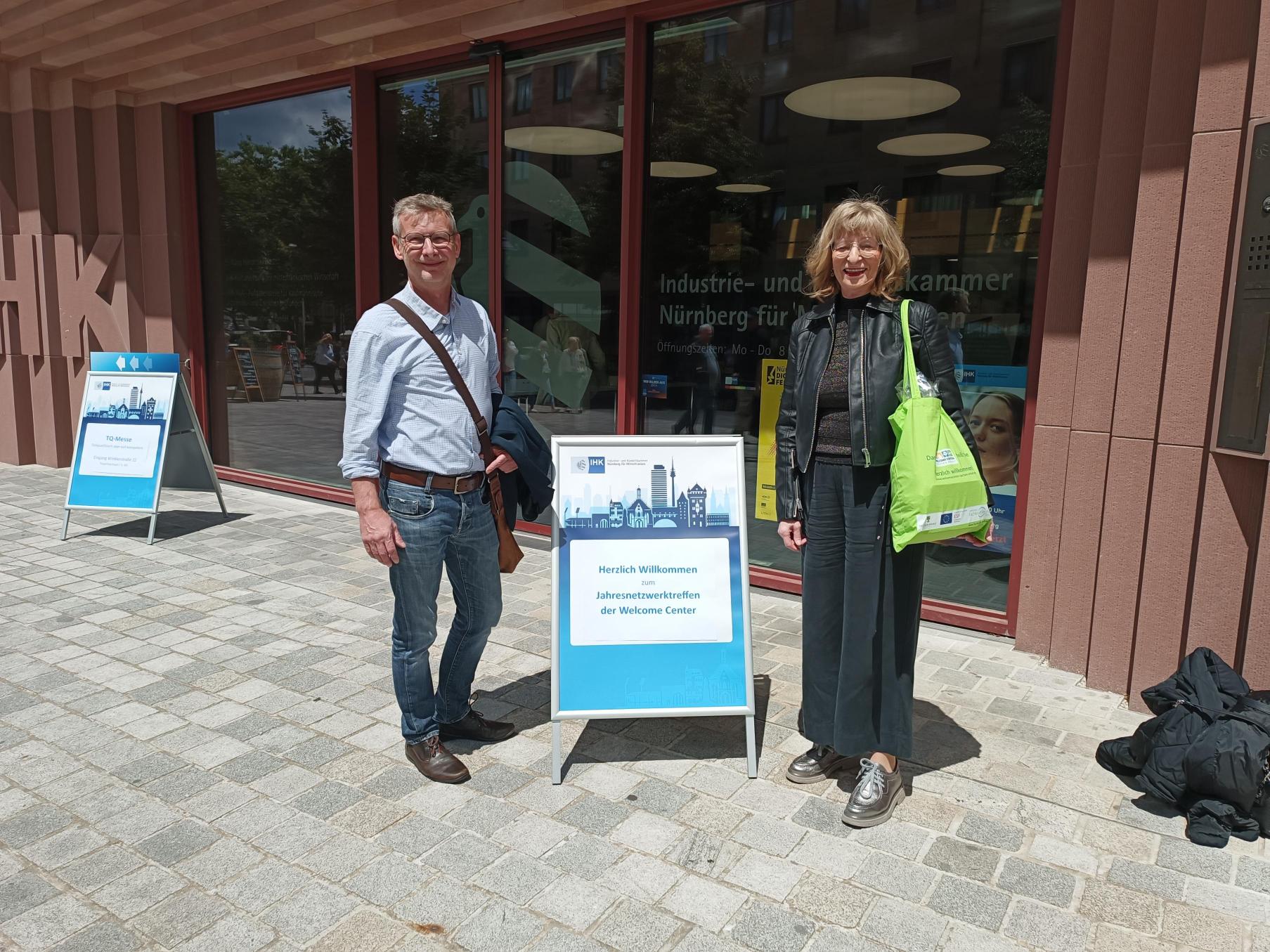 zwei Mitarbeitende des WelcomeCenters Sachsen-Anhalt vor dem Gebäude der IHK Nürnberg mit einem Aufsteller der Veranstaltung "Jahresnetzwerktreffen der Welcome Center"