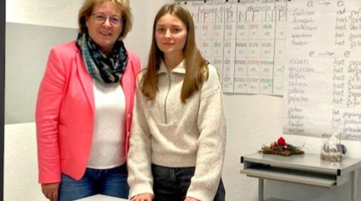 Zwei Frauen stehen an der Schulbank in einem Klassenzimmer vor einer voll beschriebenen Tafel