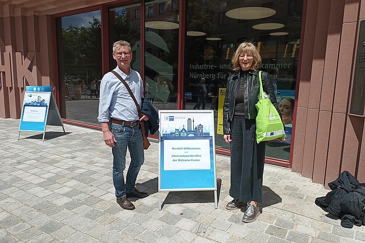 zwei Mitarbeitende des WelcomeCenters Sachsen-Anhalt vor dem Gebäude der IHK Nürnberg mit einem Aufsteller der Veranstaltung "Jahresnetzwerktreffen der Welcome Center"