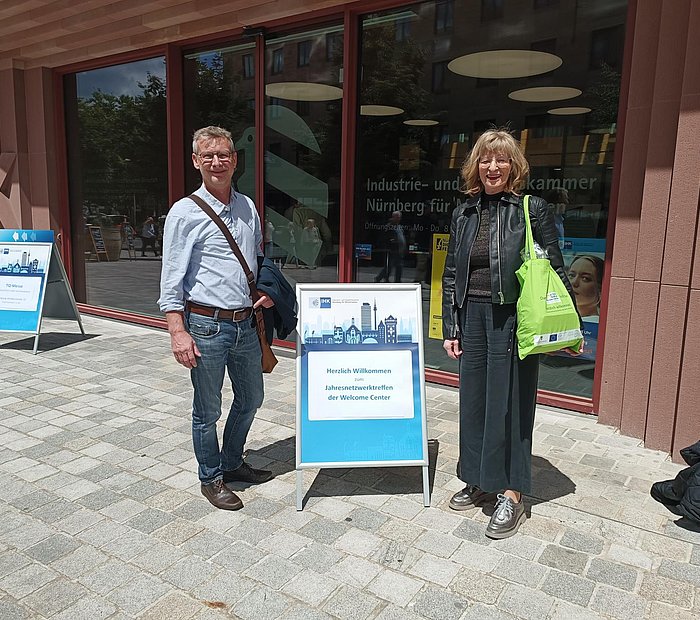 zwei Mitarbeitende des WelcomeCenters Sachsen-Anhalt vor dem Gebäude der IHK Nürnberg mit einem Aufsteller der Veranstaltung "Jahresnetzwerktreffen der Welcome Center"